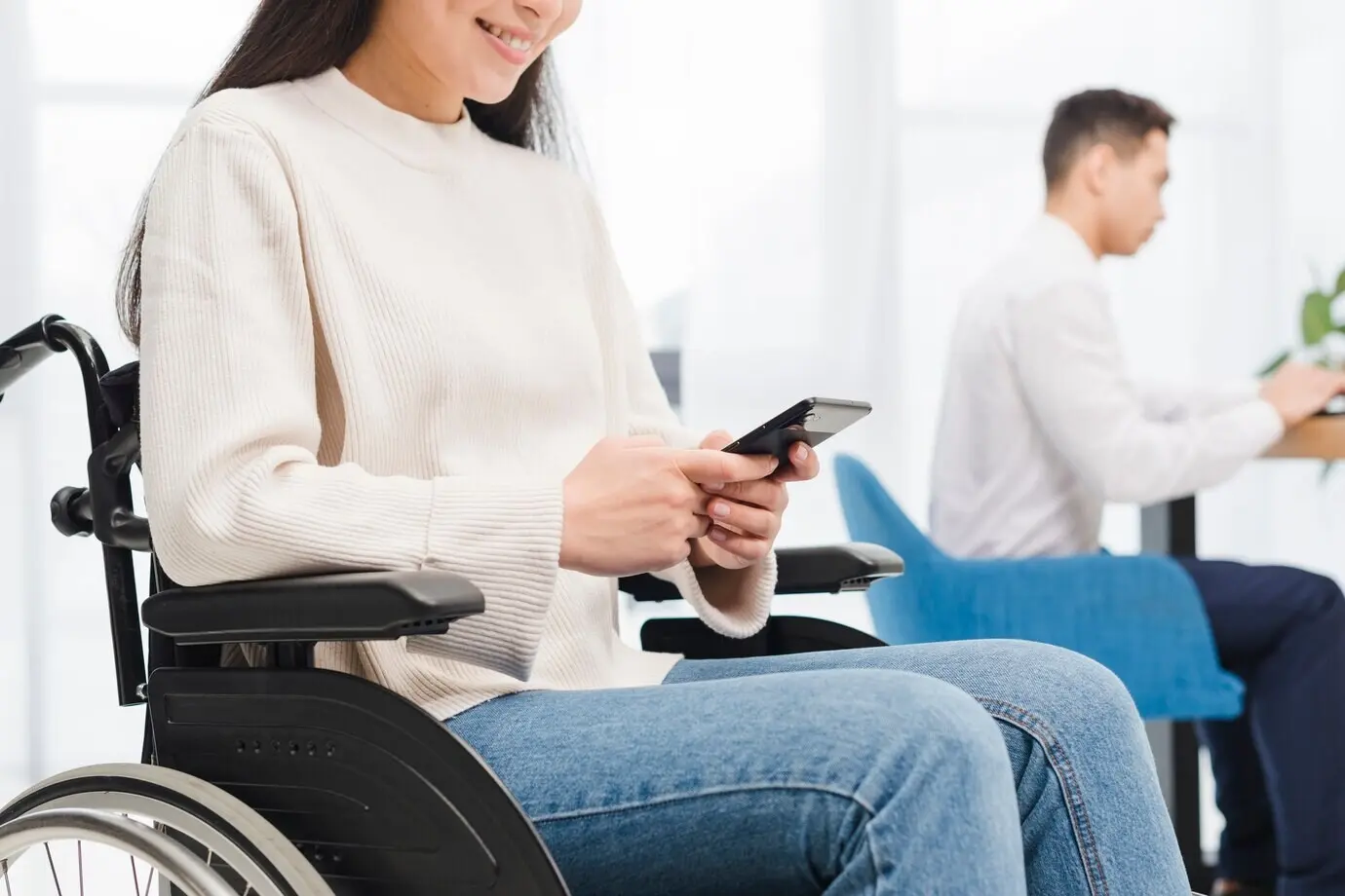 Close-up of a smiling young disabled woman in a wheelchair using a mobile phone in front of a male colleague