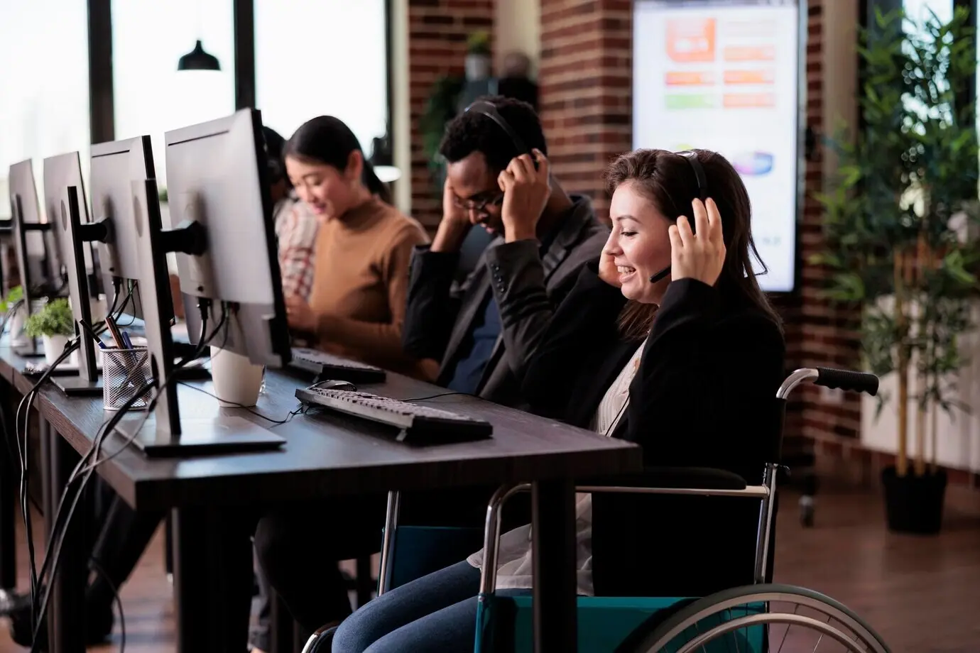 A woman working as a client support hotline operator at a call center customer service help desk in a disability-friendly office, who lives with a physical impairment and a chronic health condition and uses a wheelchair.