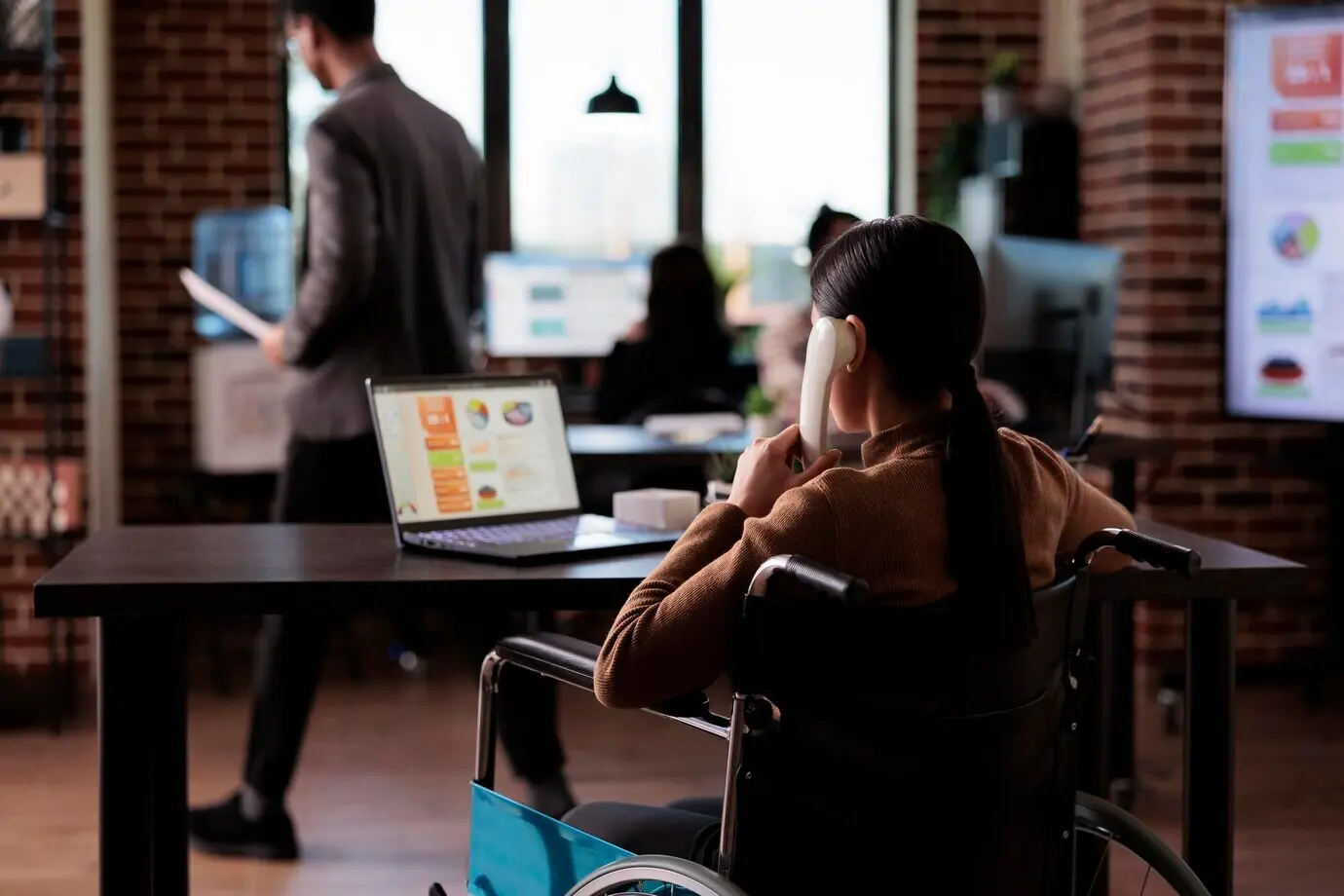A company employee with a physical impairment, who uses a wheelchair and has a chronic health condition, is speaking on an office landline during a remote phone conversation.